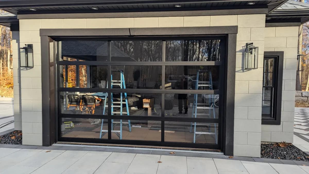 Modern full-view glass garage door with black aluminum frame on a luxury home with stone facade