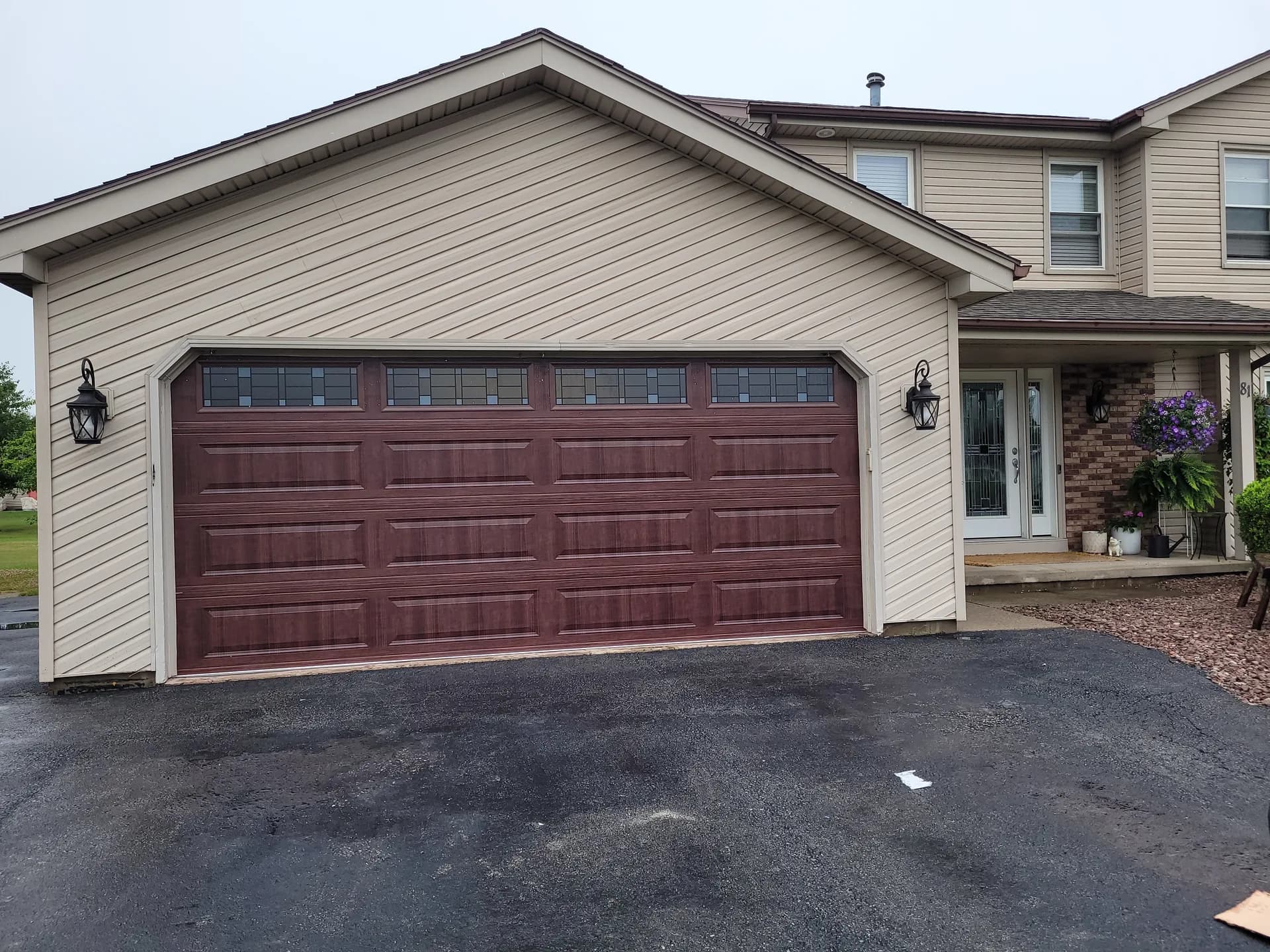 Dark walnut woodgrain garage door with leaded transom windows and coach lights on a tan home with brick accents