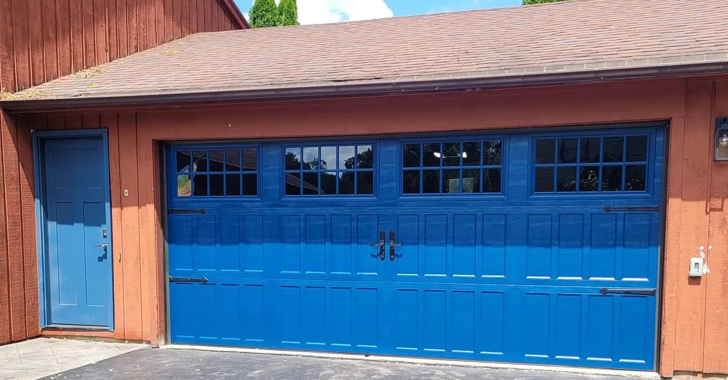 Bold blue carriage-style garage door with grid windows and decorative hardware on a red barn-style home