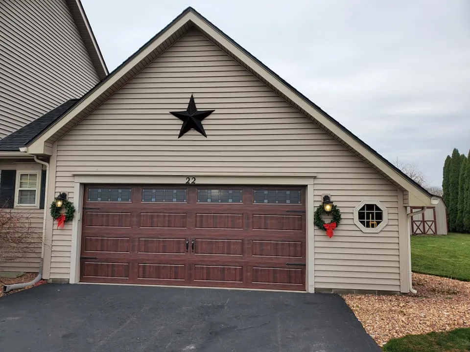Walnut woodgrain garage door with glass windows and holiday wreaths on a tan-sided home