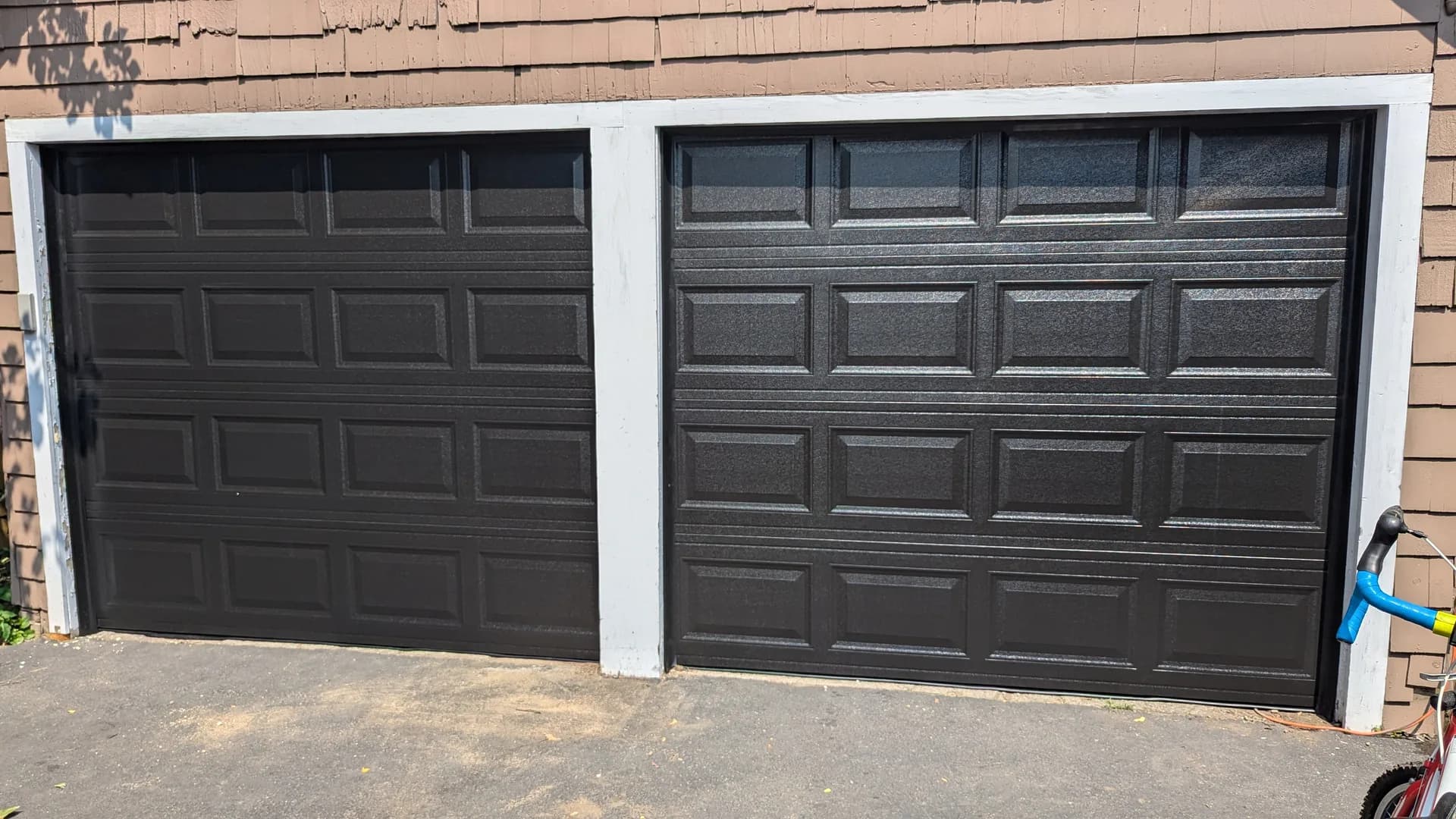 Pair of black raised-panel garage doors on a home with tan siding