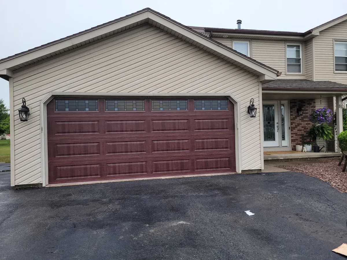 Dark walnut woodgrain garage door with leaded transom windows and coach lights on a tan home with brick accents